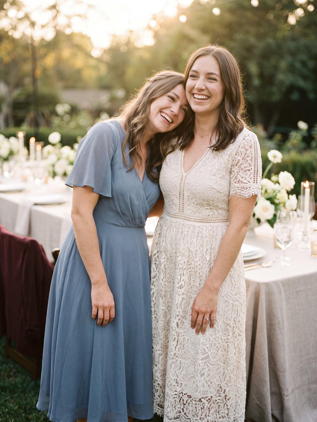 Two friends in a dusty blue chiffon midi dress and a cream lace midi dress laughing together at a garden dinner